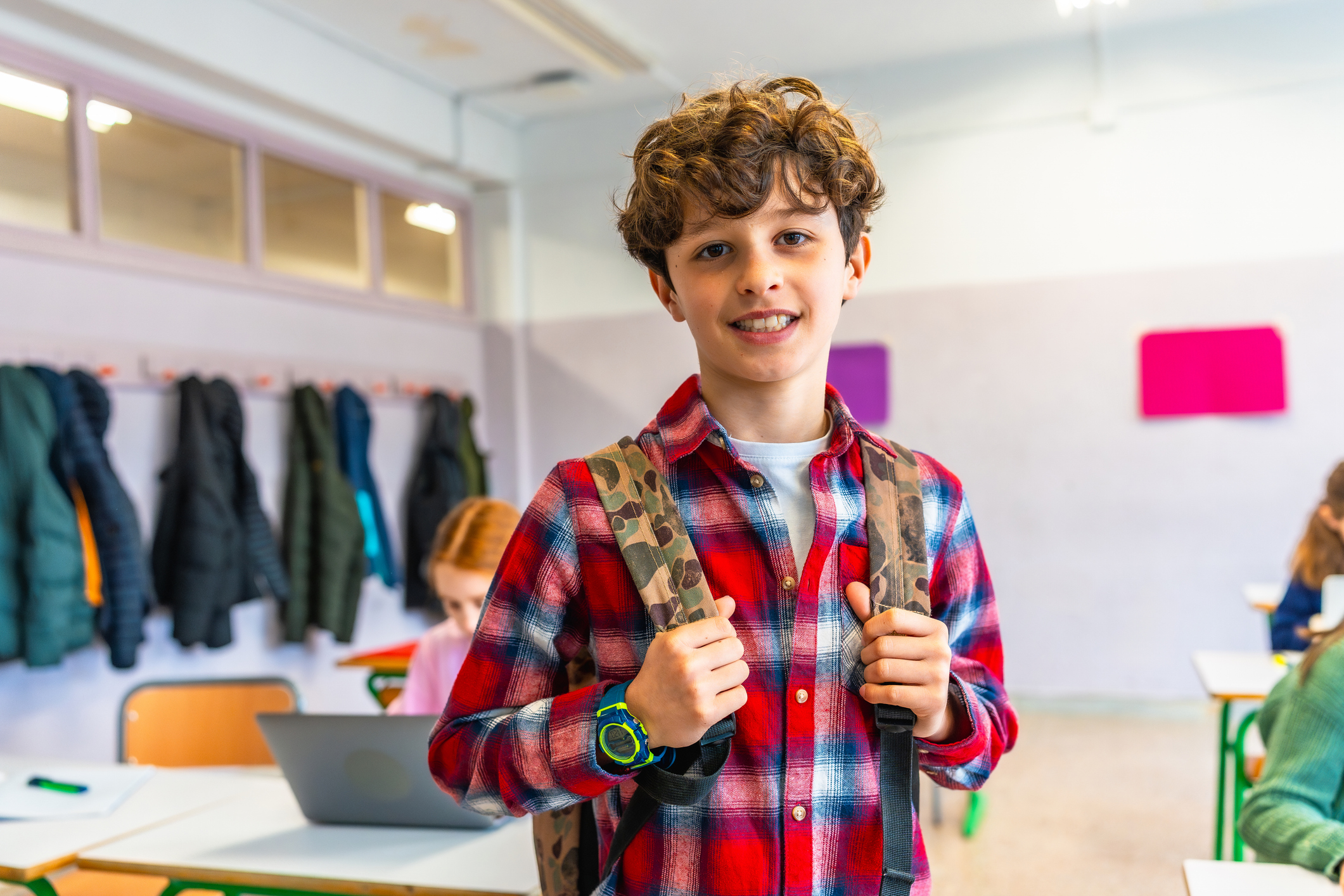 Smiling student wearing a backpack sits at a desk in a classroom, engaging with learning materials and radiating happiness